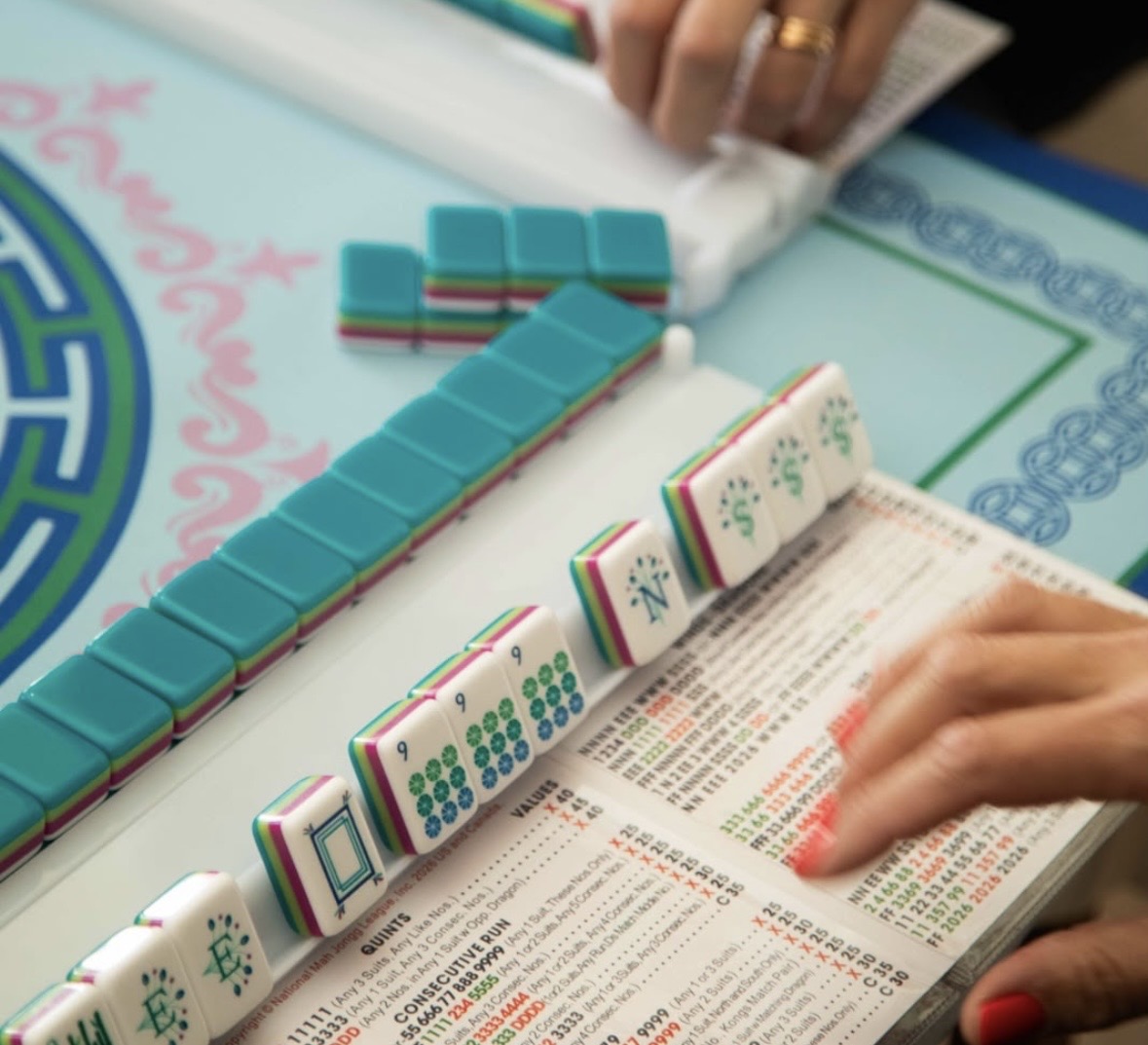 Hands playing American Mahjong with teal tiles arranged on a light blue mat beside a scorecard